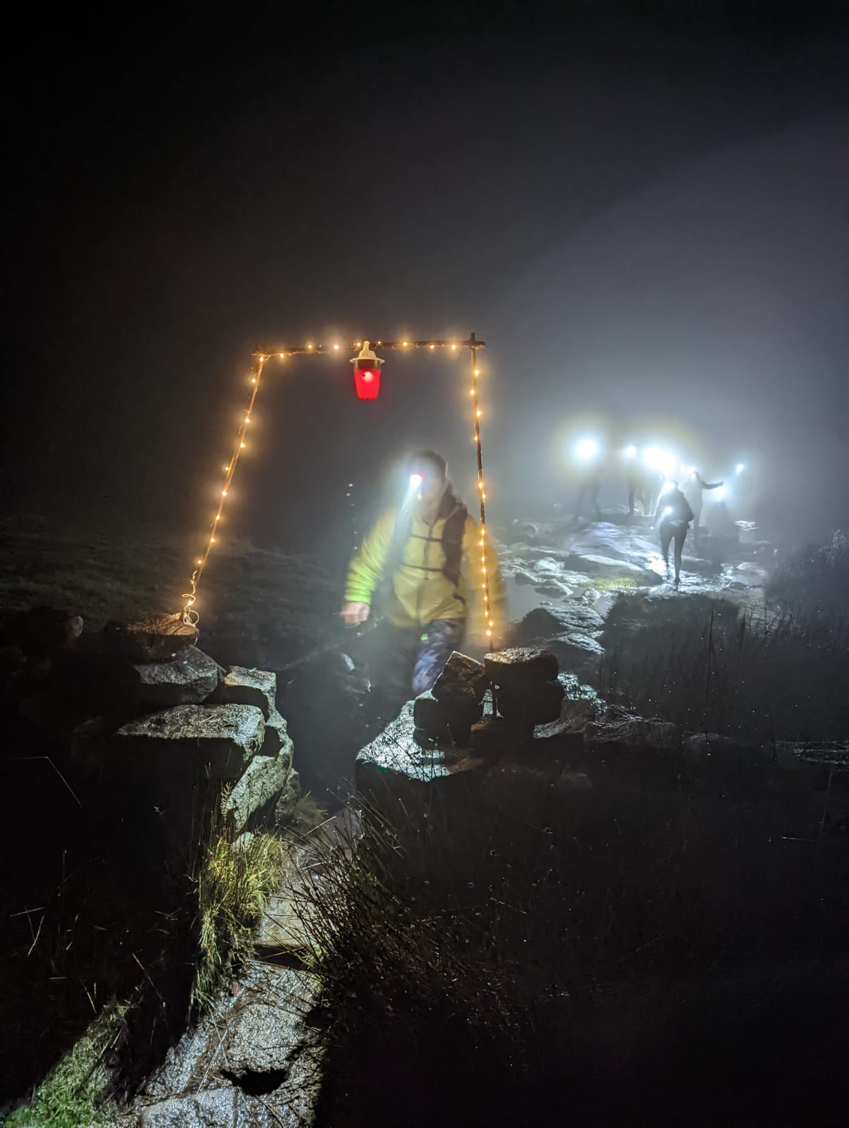 A runner in a head torch passing under a lit archway in the mist, with more head torches on the hillside behind